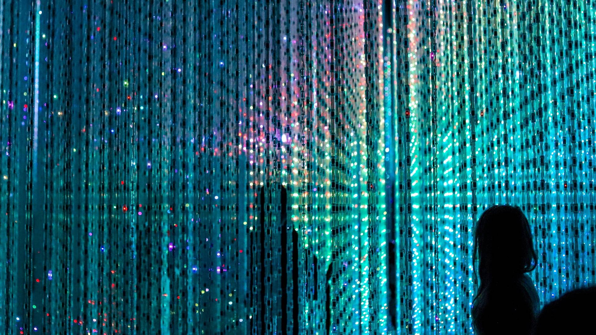 A silhouette of a person stands in front of a curtain of shimmering, multicolored LED lights creating a radiant, starburst pattern in shades of blue, green, and purple.