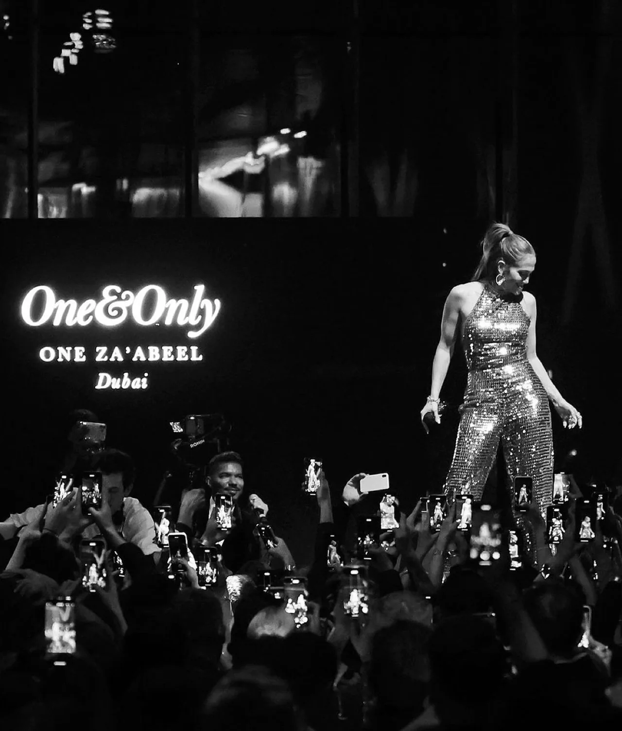 A woman in a sparkling outfit performs on stage at the One&Only One Za’abeel Dubai event, as a large crowd takes photos and videos with their phones. The image is in black and white.