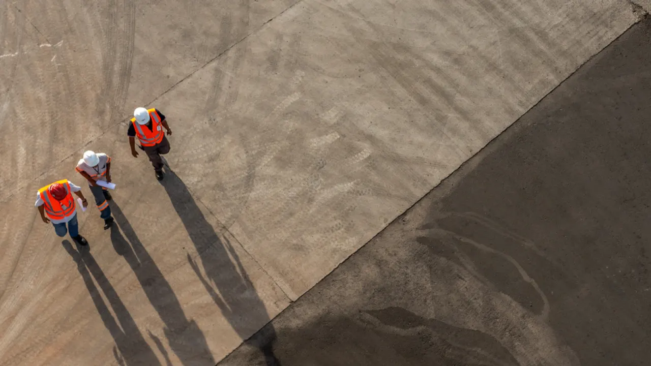 Three construction workers in safety vests and helmets stand on a large concrete surface, casting long shadows, with a dark asphalt area visible to the right.