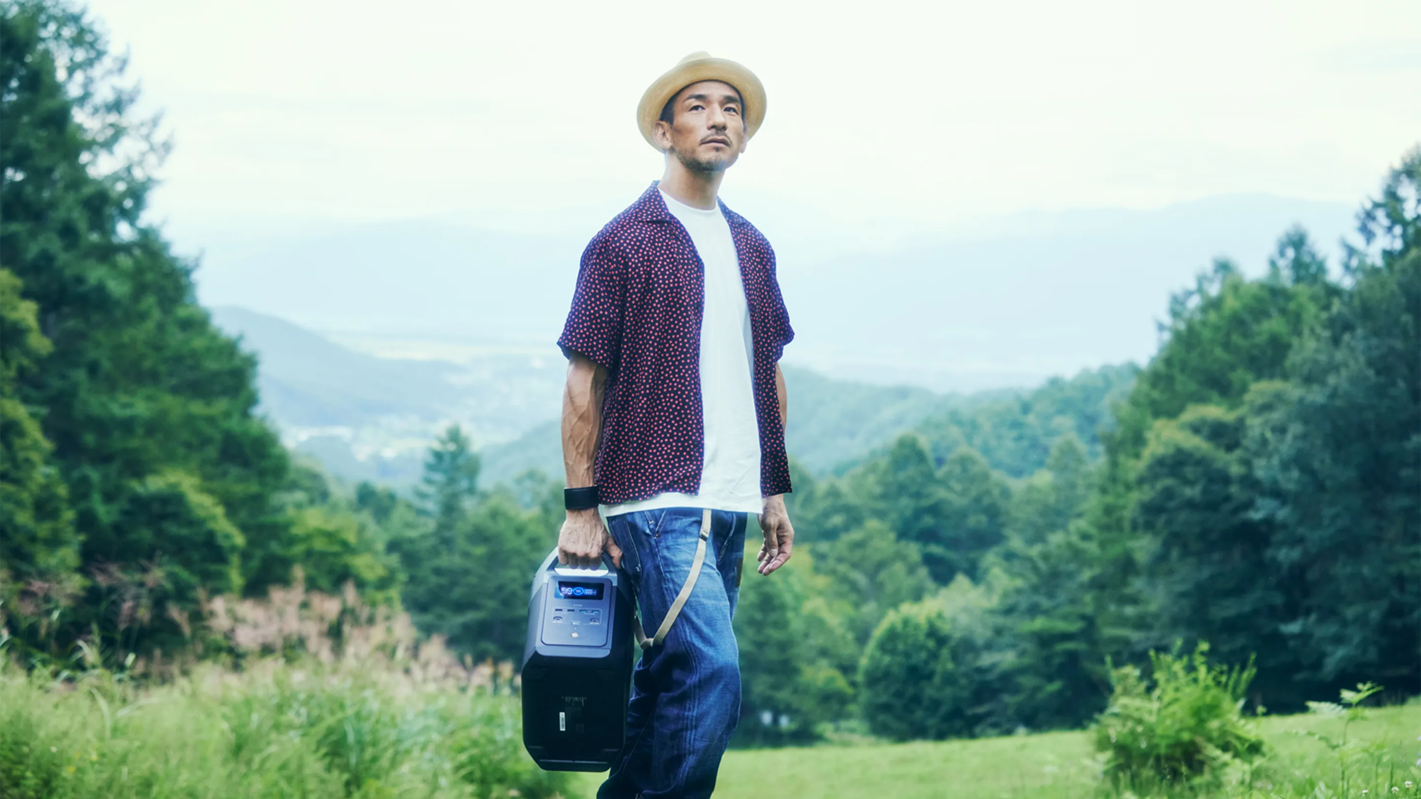 A man wearing a hat and casual clothes stands in a grassy clearing, holding a portable speaker, with green trees and distant hills in the background.