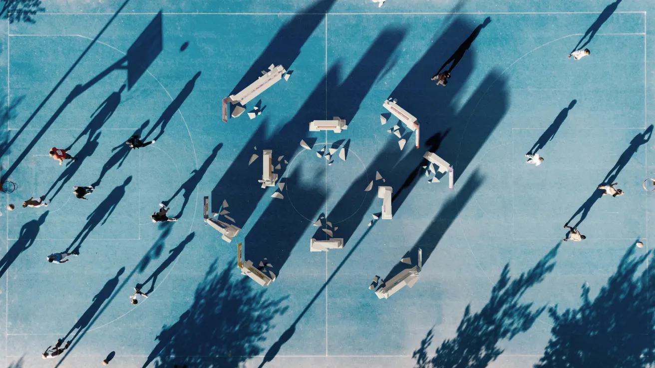 Aerial view of people walking on a blue sports court with rectangular objects casting long shadows. The scene includes scattered figures and tree shadows, creating an abstract pattern on the surface.