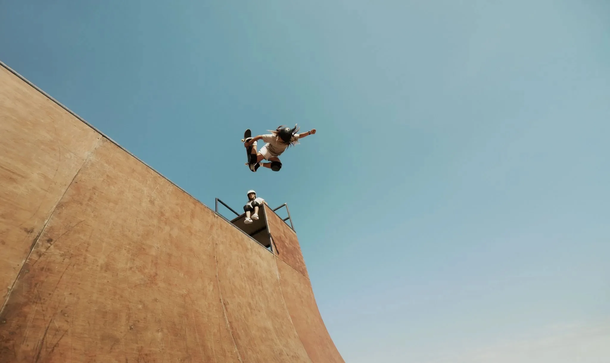 A skateboarder performs a high aerial trick above a large wooden ramp, while another person sits on the edge watching, all against a clear blue sky.