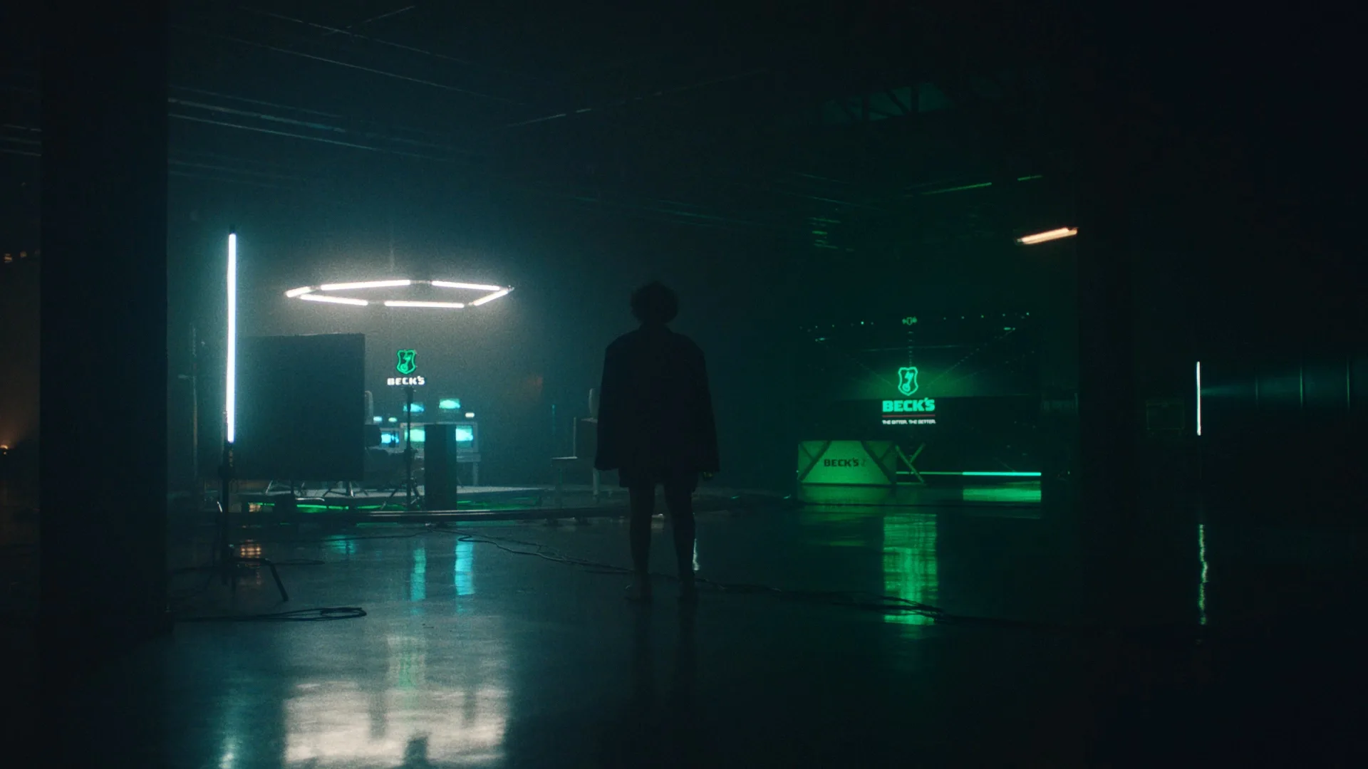 A person stands in a dark, futuristic room lit by neon green and blue lights, with illuminated screens and equipment reflecting on the shiny floor. The BECK’S logo is visible in the background.