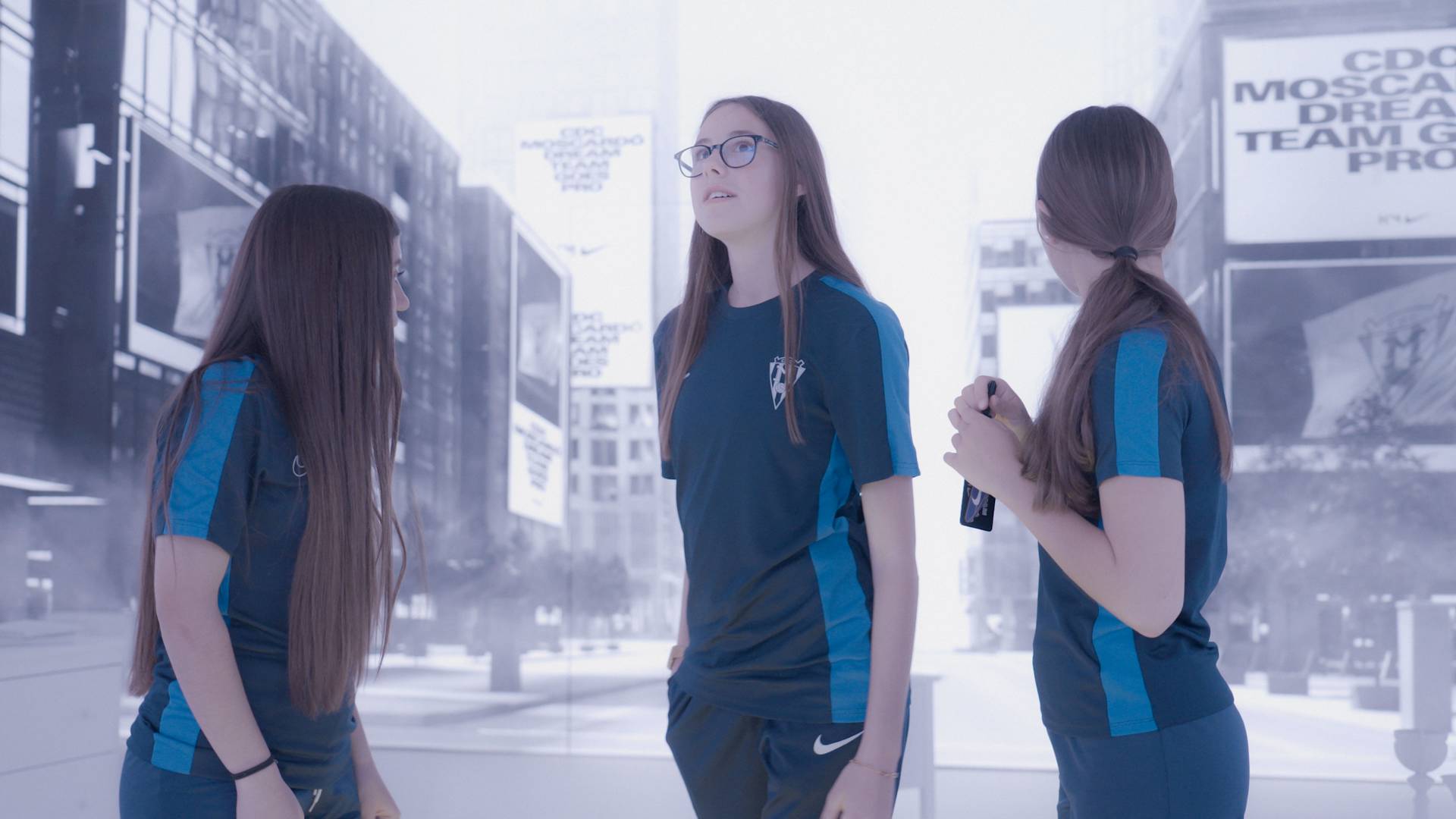 Three young women in matching blue sports uniforms stand together indoors, looking in different directions. The background displays a cityscape with large text on buildings, giving a modern, urban feel.