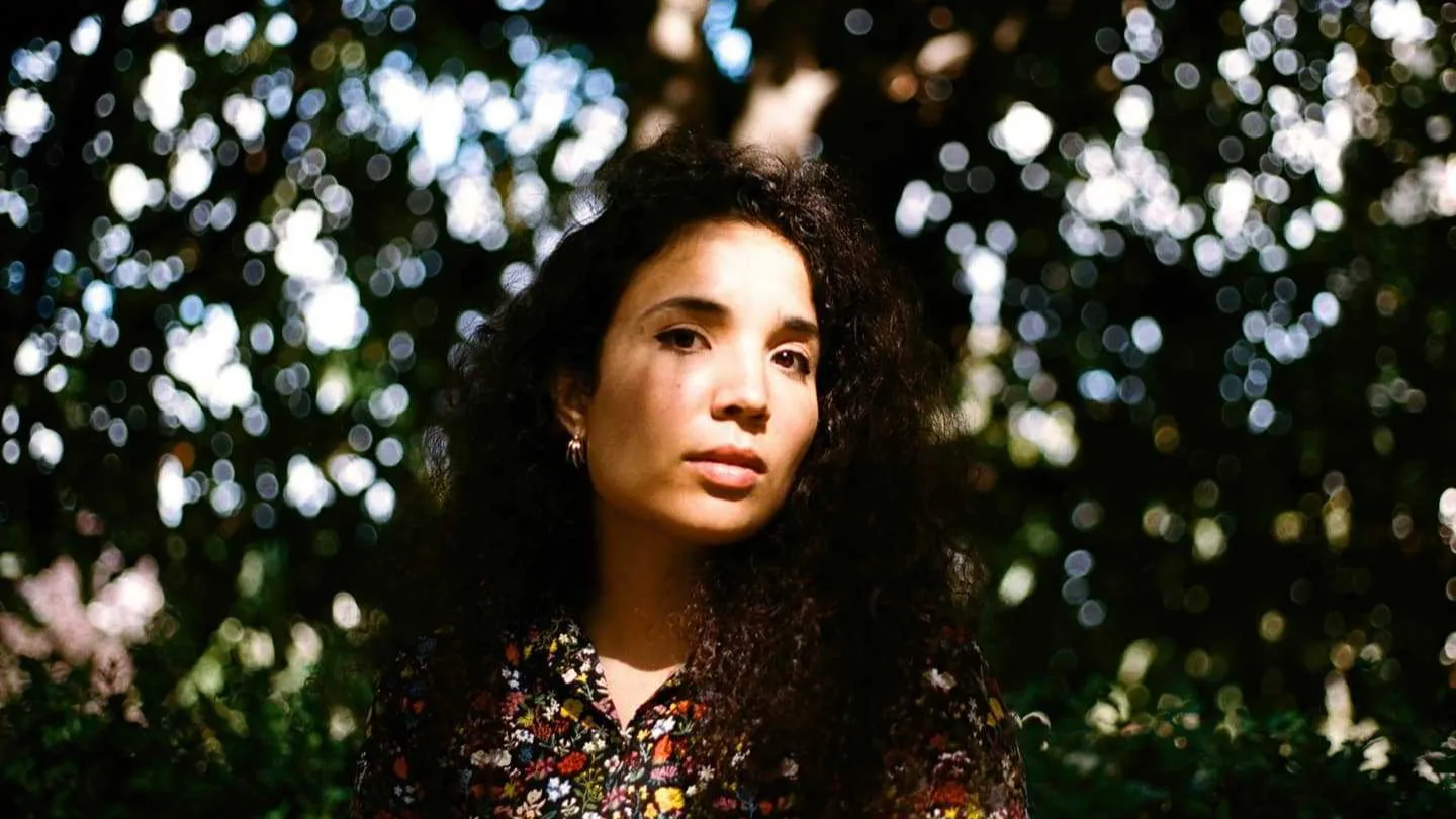 A woman with long, curly dark hair and a floral shirt stands outdoors, looking at the camera. Sunlight filters through trees behind her, creating a soft, bokeh effect.