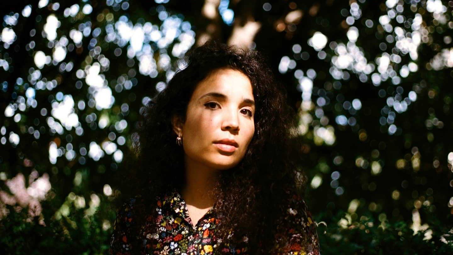 A woman with long, curly dark hair and a floral shirt stands outdoors, looking at the camera. Sunlight filters through trees behind her, creating a soft, bokeh effect.