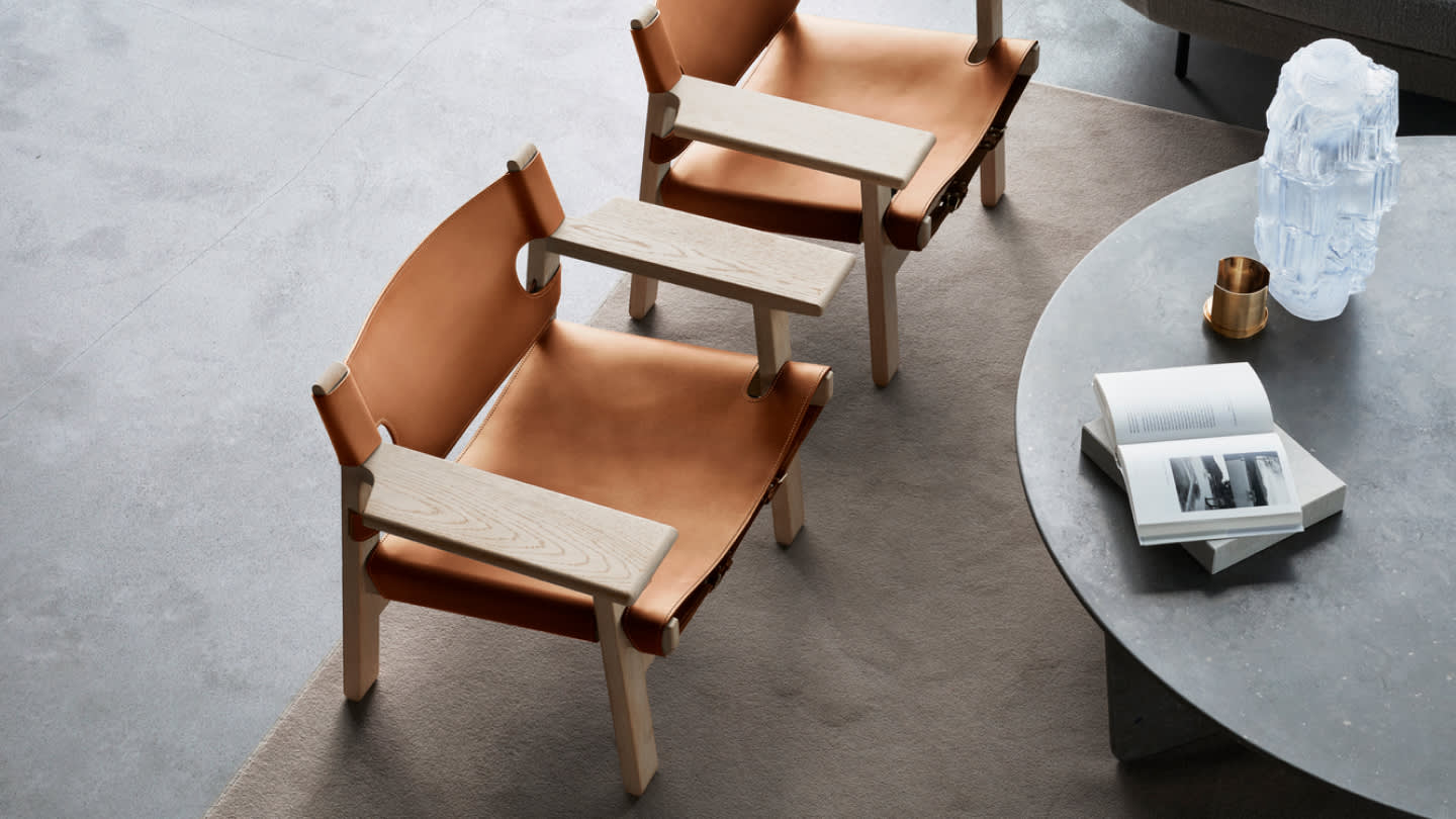 Two modern tan leather and light wood lounge chairs are placed next to a round gray coffee table with an open book, a gold cup, and a sculptural glass object on top, all on a gray rug in a minimalist living room.