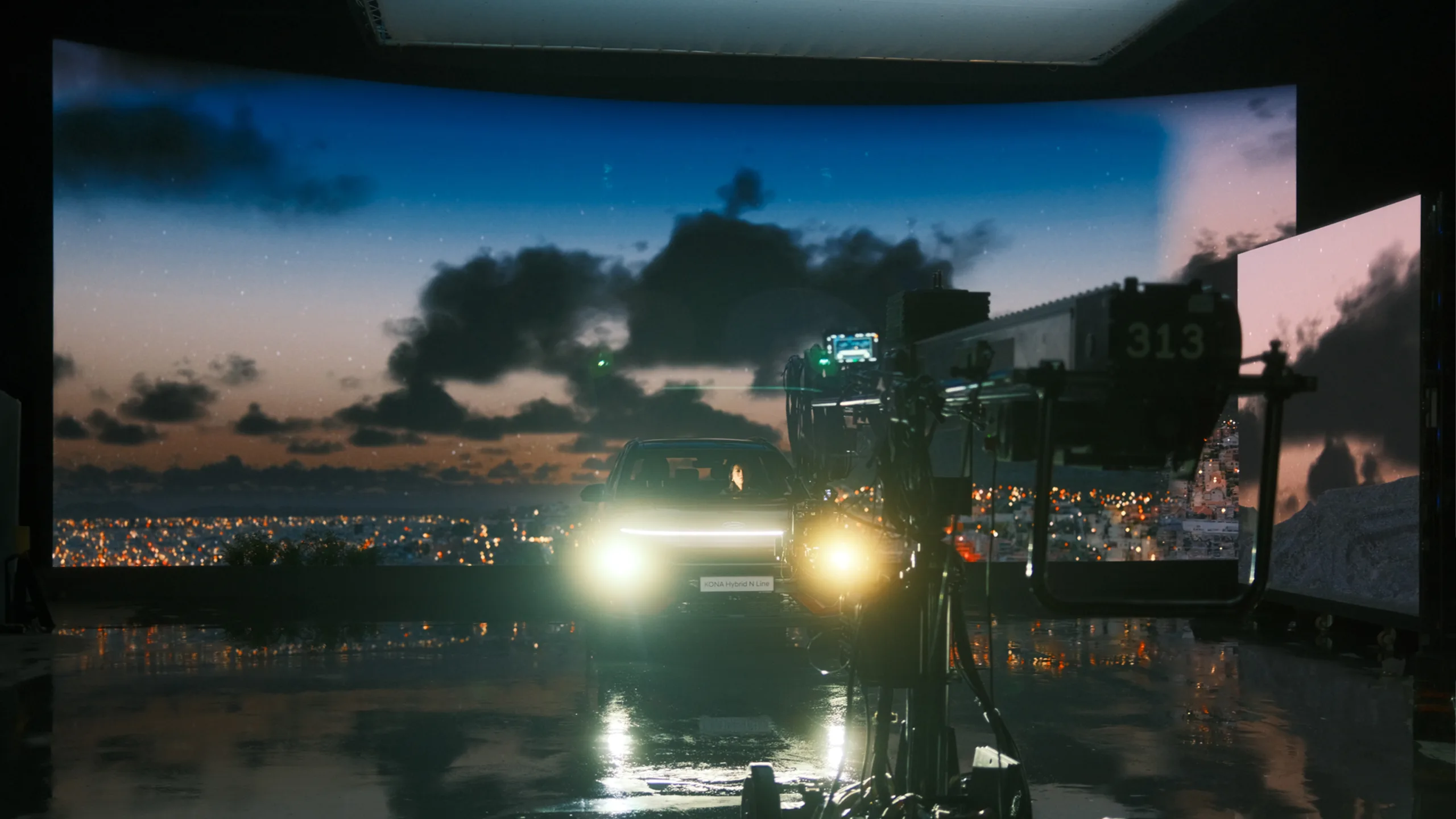 A car with headlights on is parked in a studio, surrounded by large screens displaying a cityscape at dusk and a crane camera in the foreground, reflecting on the shiny floor.