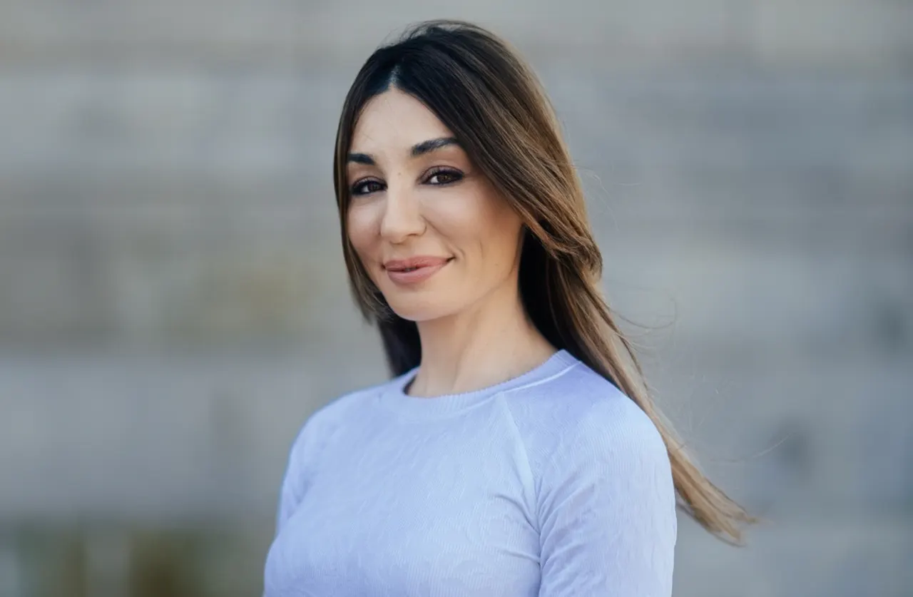 A woman with long brown hair and a light blue top smiles slightly while standing outdoors in front of a blurred stone wall.