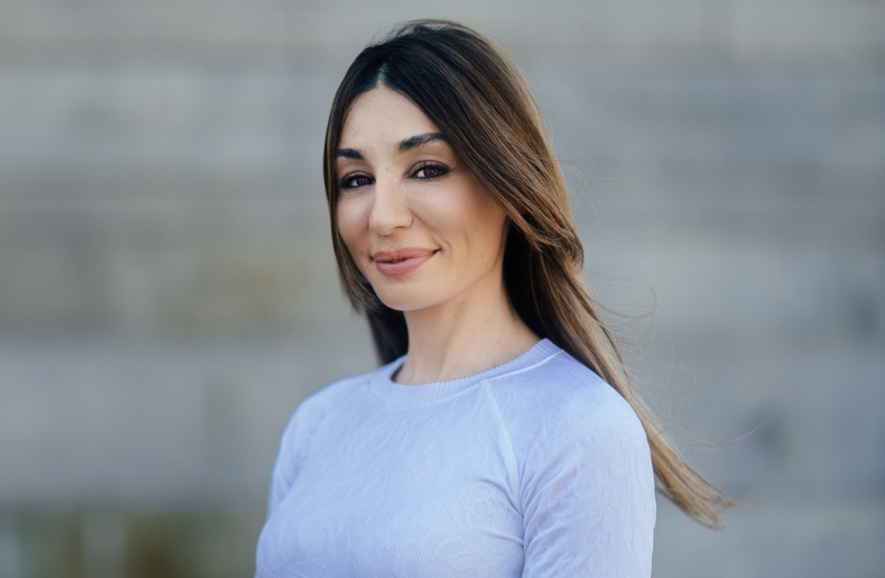 A woman with long brown hair and a light blue top smiles slightly while standing outdoors in front of a blurred stone wall.