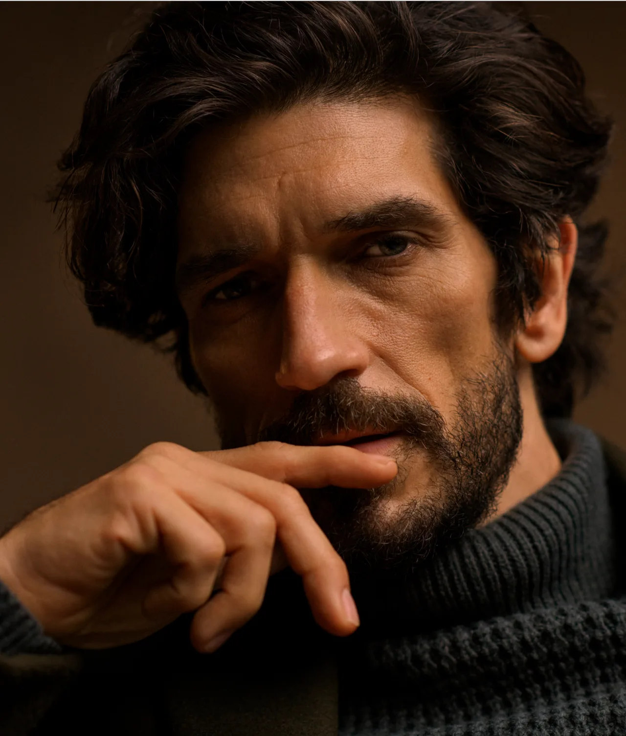 A man with wavy dark hair and a beard gazes thoughtfully at the camera, his hand resting near his lips. He wears a dark, textured turtleneck sweater against a plain, warm-toned background.