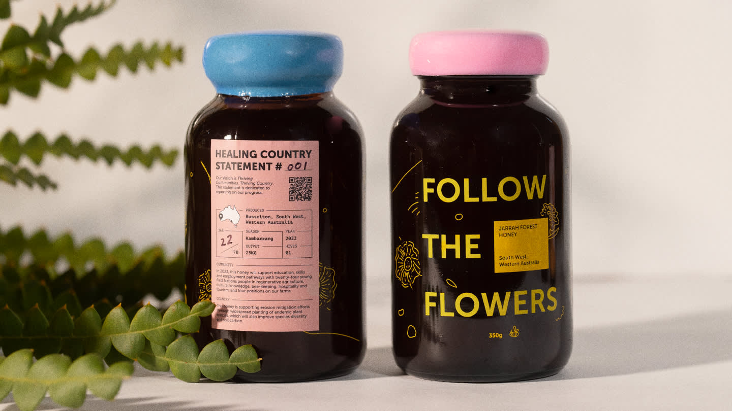 Two glass jars of honey with blue and pink lids are shown. One jar displays product information, while the other has “FOLLOW THE FLOWERS” in yellow text. Fern leaves frame the jars on a light surface.