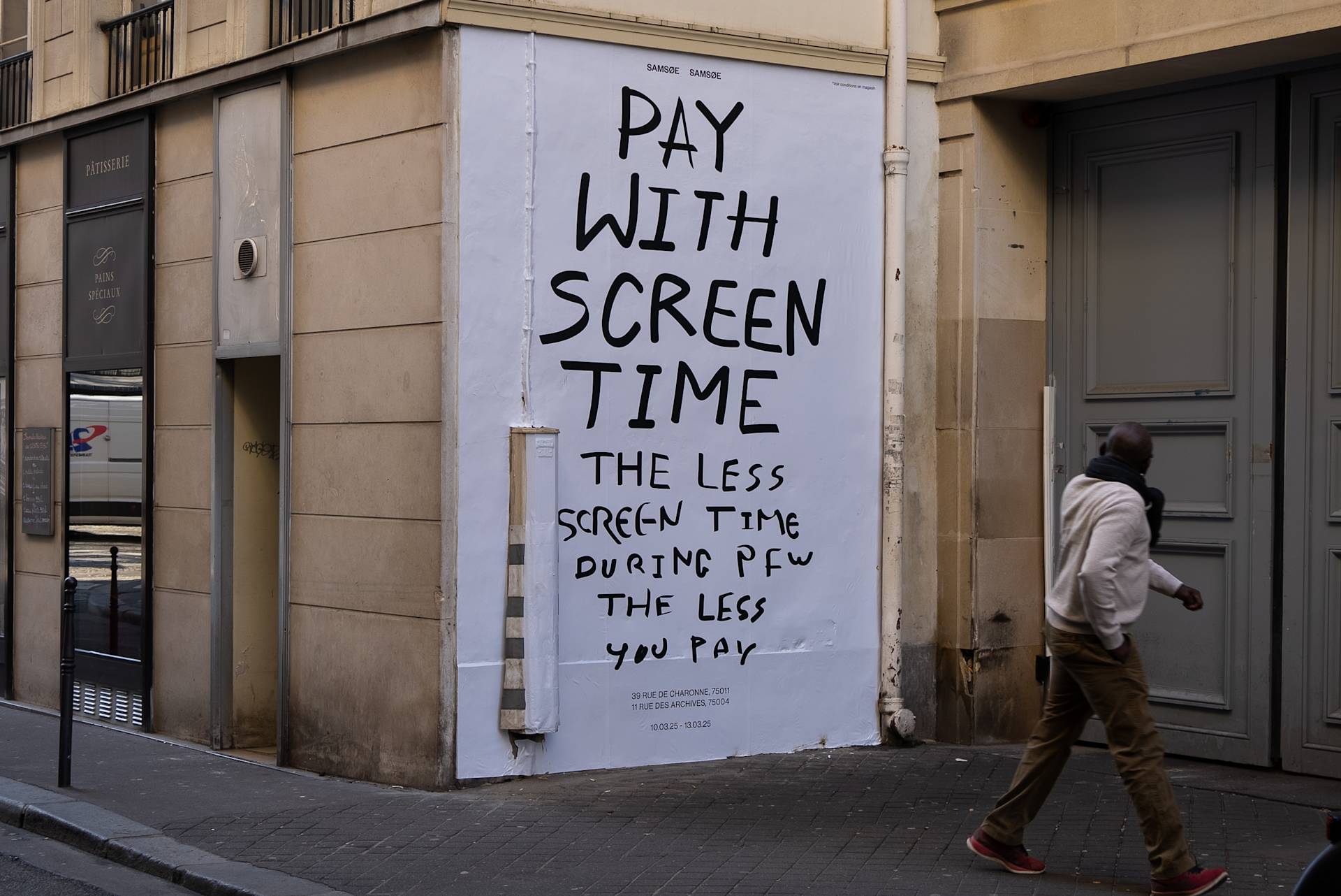 A man walks past a large white poster on a city street wall that reads, Pay with screen time. The less screen time during pay, the less you pay, in handwritten black letters.