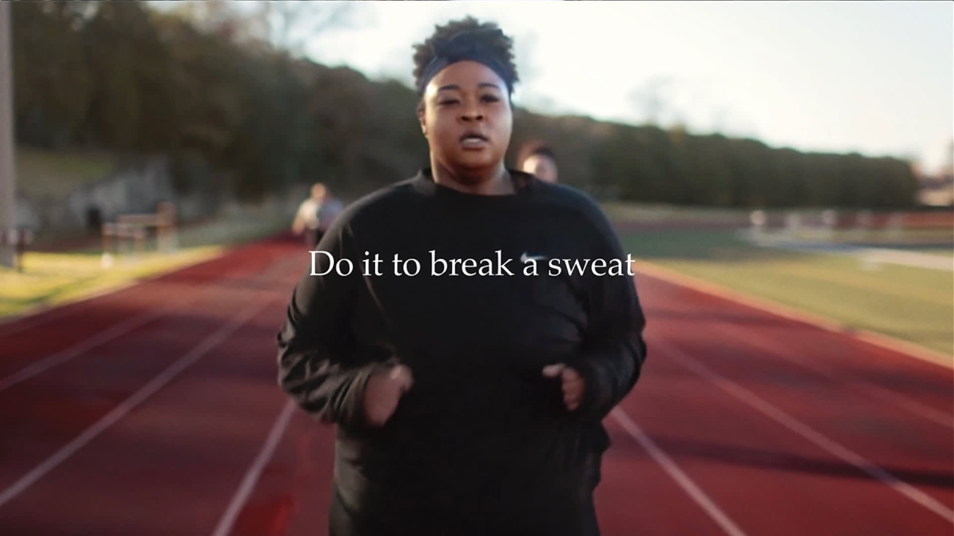 A person in a black sweatshirt jogs on an outdoor track, with blurred runners in the background. Text over the image reads, “Do it to break a sweat.”.