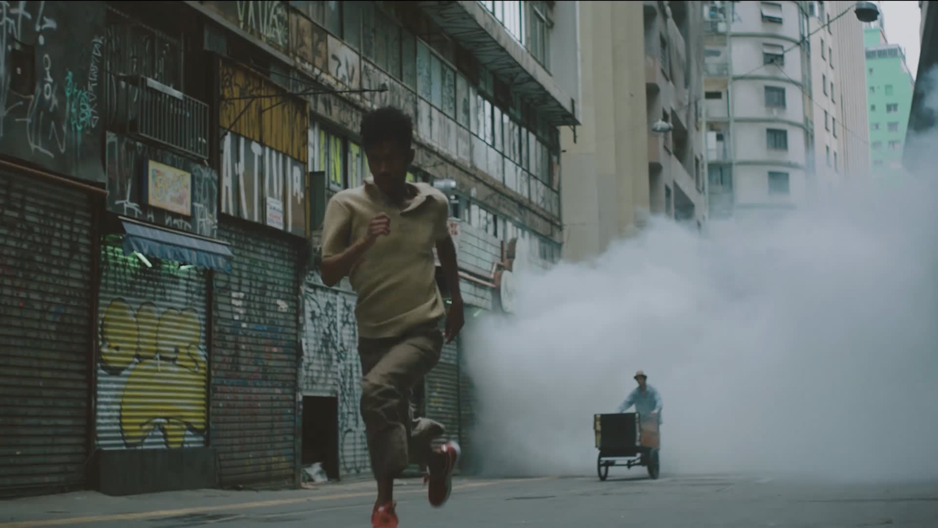 A man runs down a graffiti-covered urban street while a person in the background pushes a cart emitting thick white smoke. The atmosphere appears tense and dramatic.