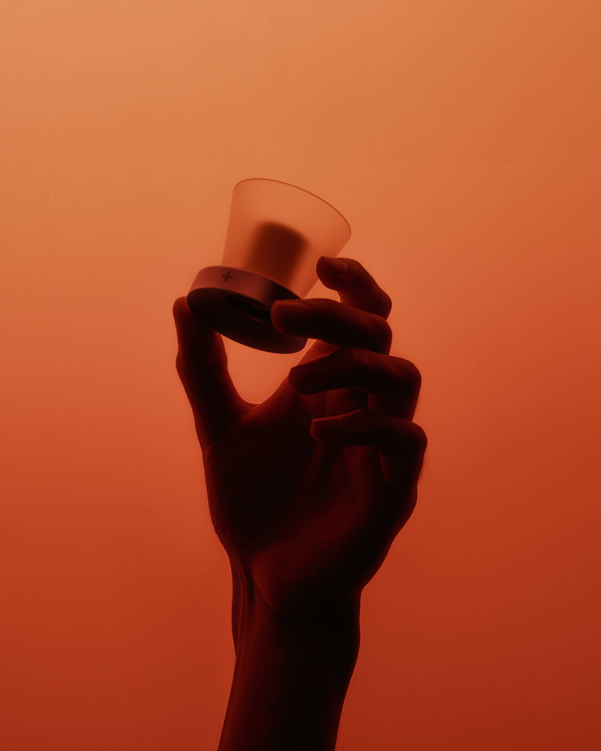A silhouetted hand holds a small, frosted glass cup against an orange background, creating a dramatic and minimalistic scene.