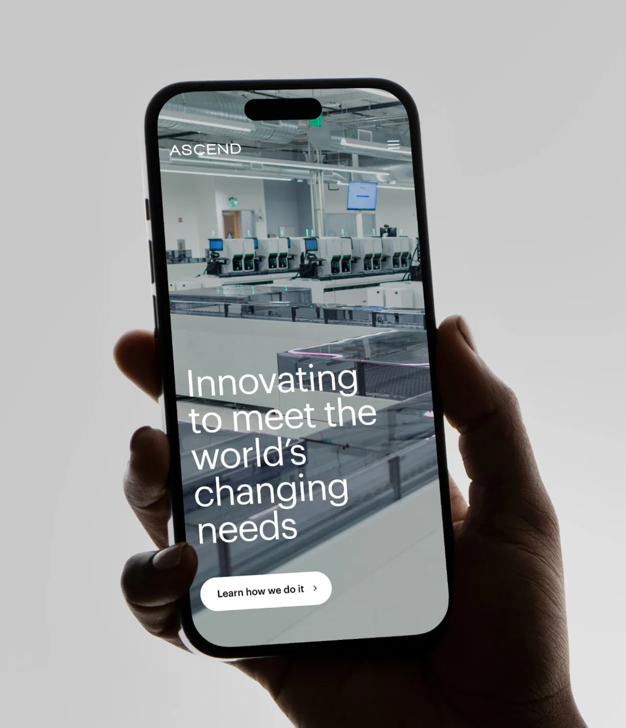 A hand holds a smartphone displaying a website with a modern factory image and the text: Innovating to meet the world’s changing needs. A button below reads Learn how we do it. The screen header says ASCEND.