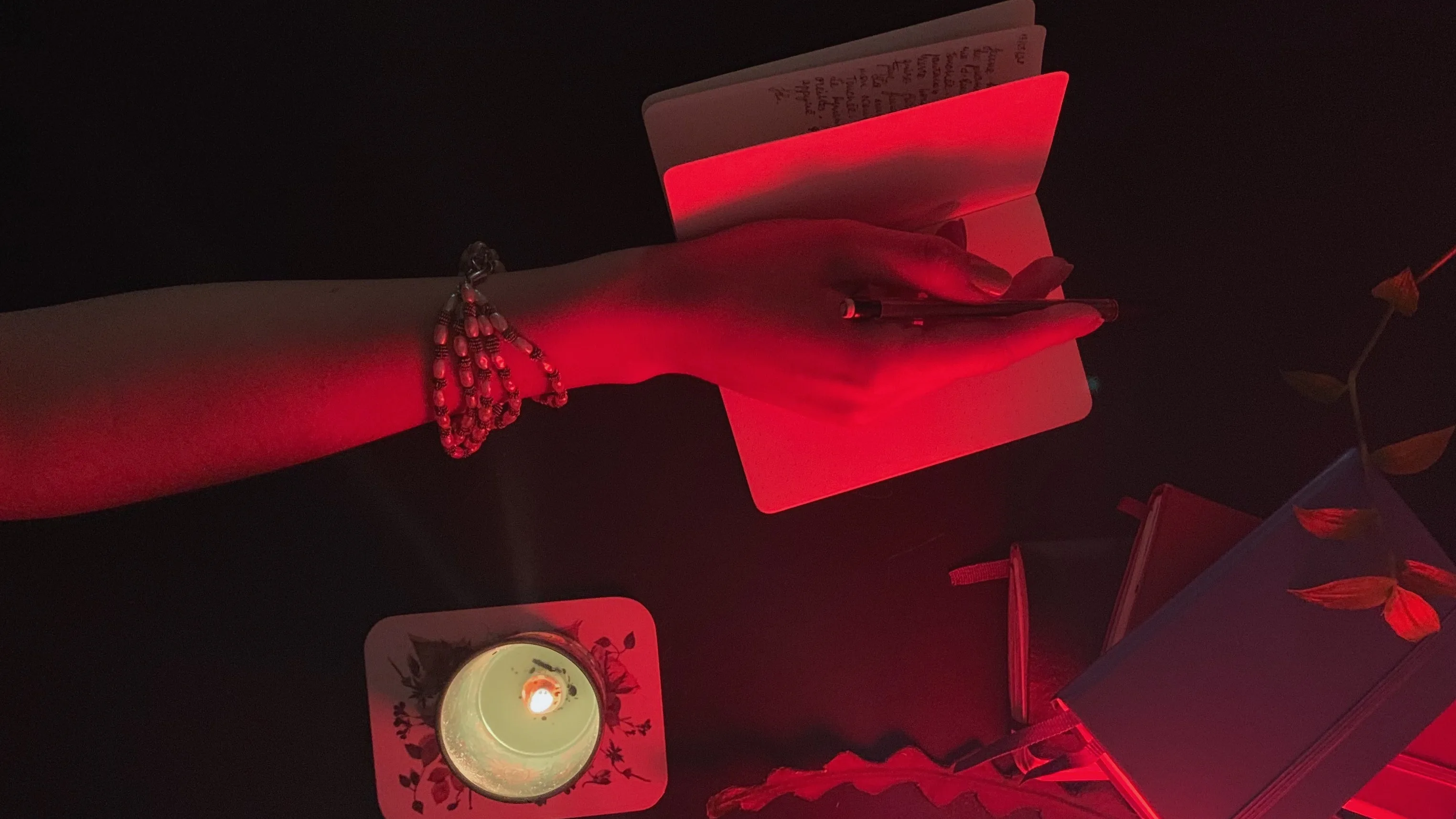 A hand wearing bracelets writes in a notebook under red light, with a lit candle on a floral coaster nearby and closed notebooks and leaves on a dark surface.