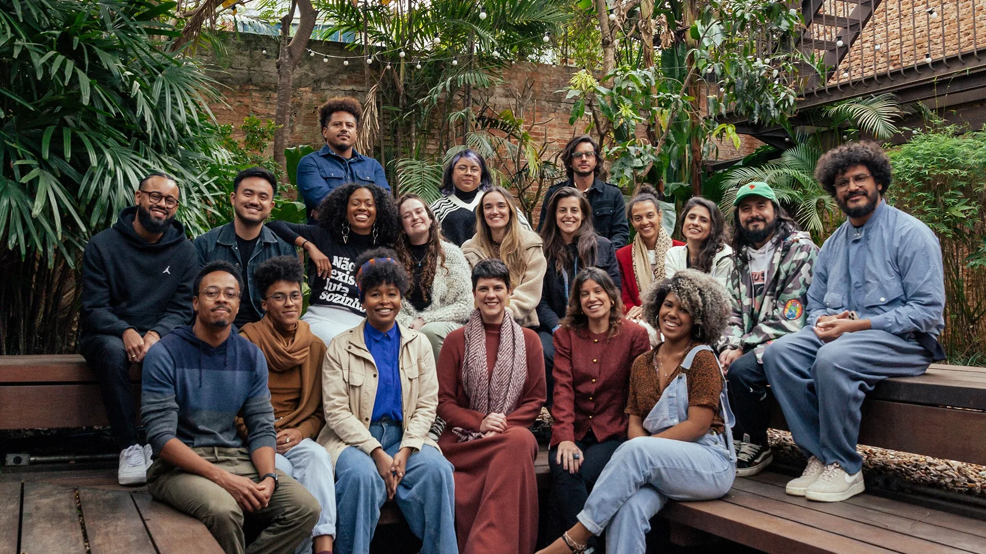 A diverse group of twenty people sit and stand on wooden benches outdoors, surrounded by lush green plants. They are smiling and casually dressed, posing together for a group photo.
