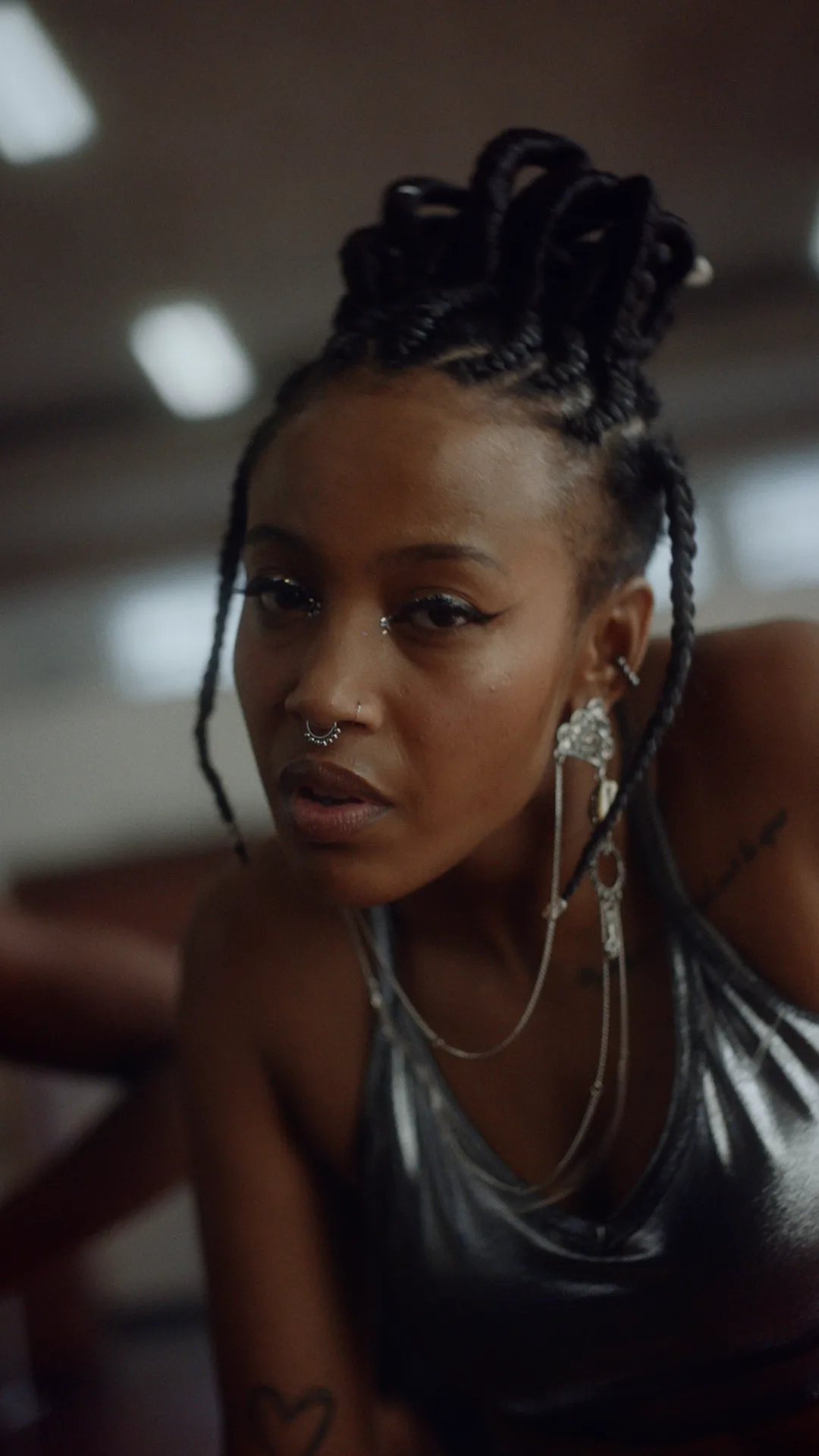 A person with braided hair, nose and septum piercings, and silver jewelry wears a shiny silver top, looking directly at the camera with a confident expression indoors.