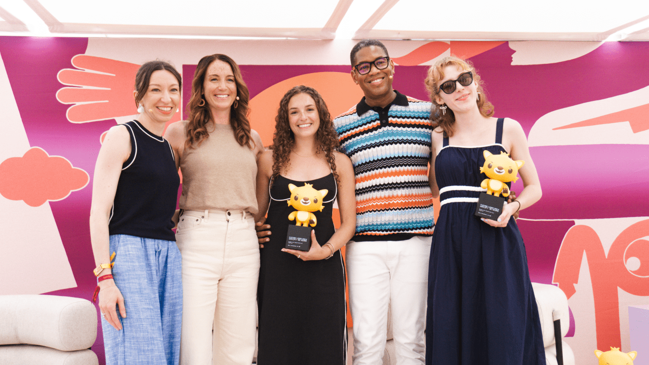 Five people stand smiling in front of a colorful mural. Two of them hold yellow cartoonish trophies. The group appears happy and celebratory, dressed in casual and semi-formal attire.