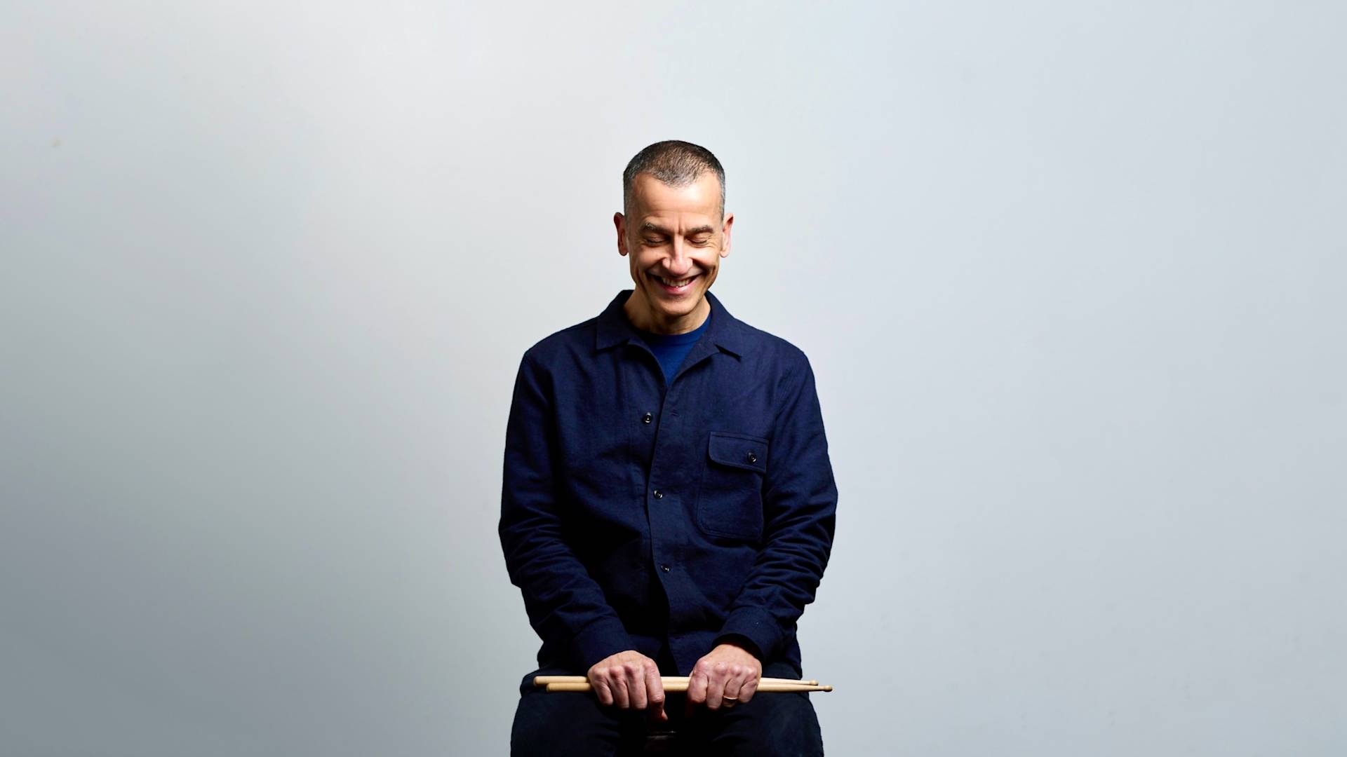 A person with short hair, wearing a dark blue jacket and dark pants, sits on a stool against a plain light background, smiling and looking down while holding a pair of drumsticks across their lap.
