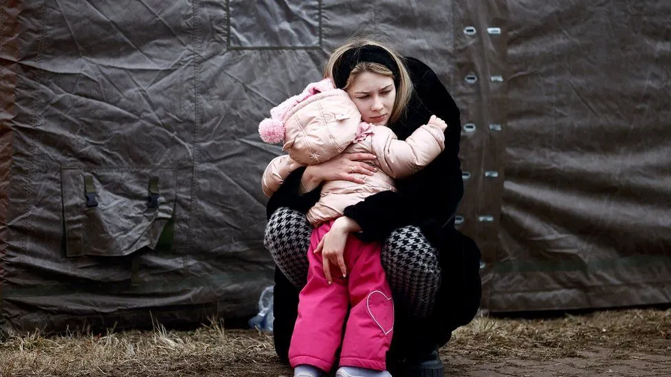 A woman kneels down and hugs a young child dressed in a pink coat and hat. They are outside, near a tent, and both appear somber. The ground is bare and the atmosphere looks cold.