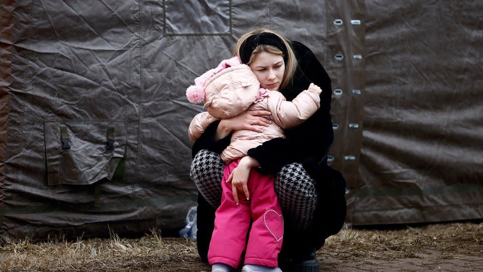 A woman kneels down and hugs a young child dressed in a pink coat and hat. They are outside, near a tent, and both appear somber. The ground is bare and the atmosphere looks cold.
