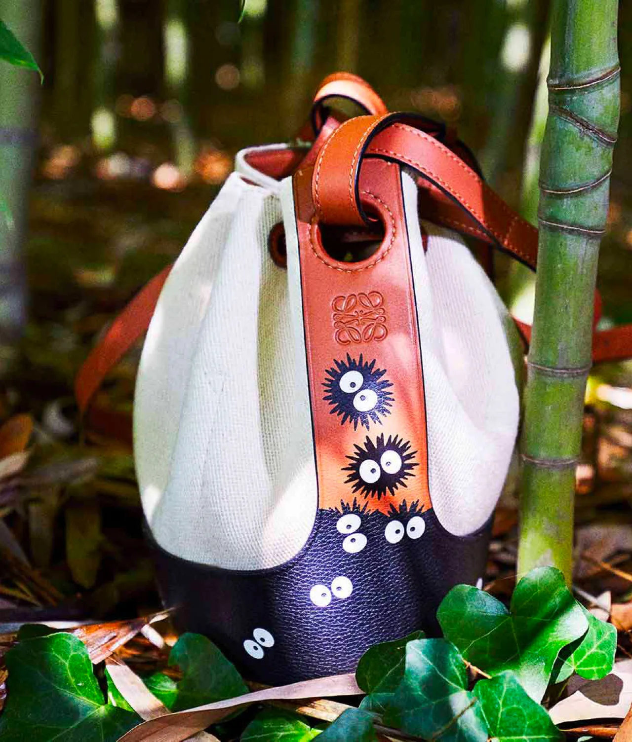 A canvas and leather bucket bag with brown handles, featuring playful black and white soot sprite designs, sits on the forest floor among green leaves and bamboo stalks.
