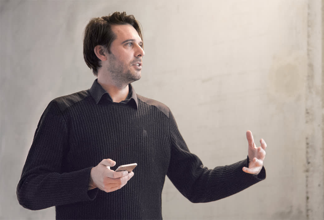A man with dark hair wearing a black sweater holds a smartphone in one hand and gestures with the other while speaking in front of a plain, light-colored wall.