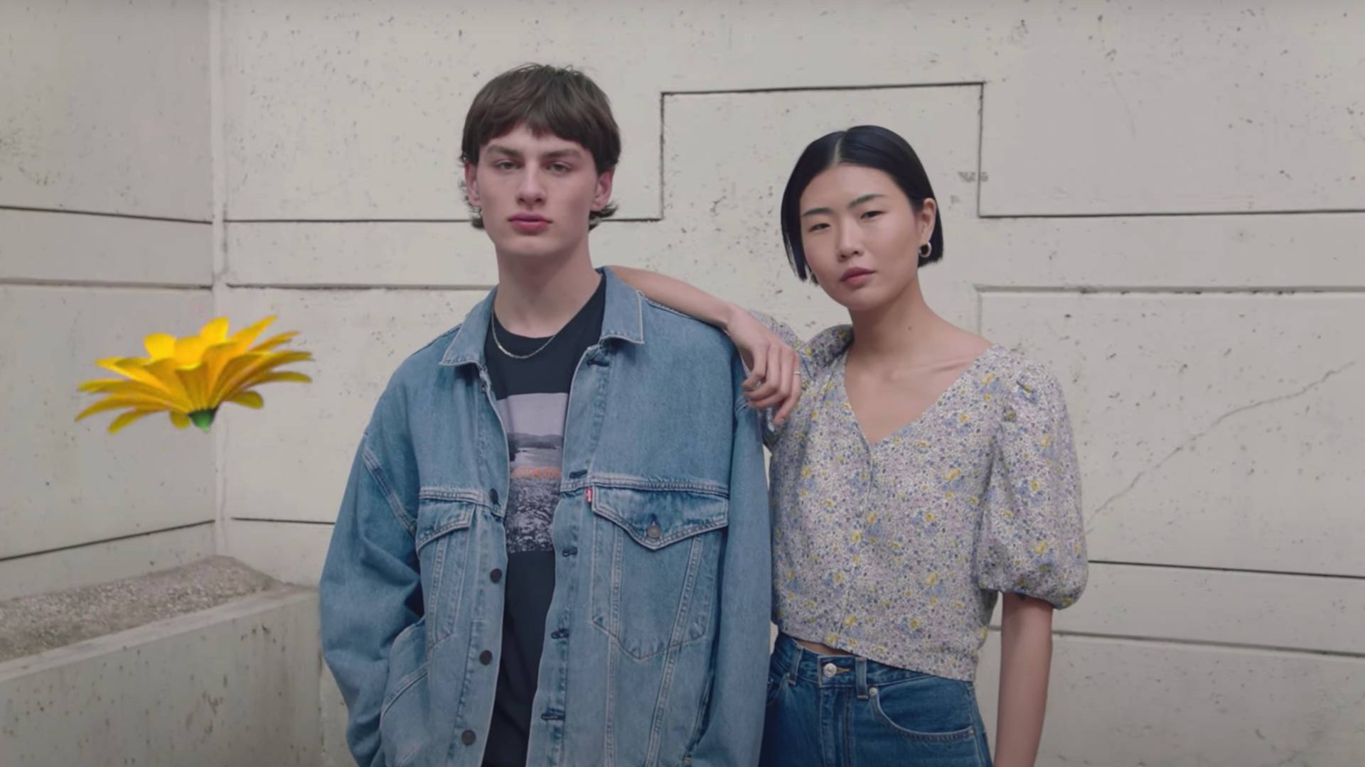 Two people stand side by side against a textured wall. One wears a denim jacket and the other a floral blouse with puff sleeves. The person in the blouse rests their arm on the others shoulder. A yellow flower floats nearby.