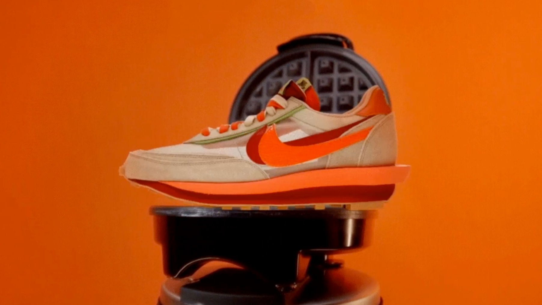 A modern beige and orange sneaker with a large orange Nike swoosh is placed on a black waffle maker against a solid bright orange background.