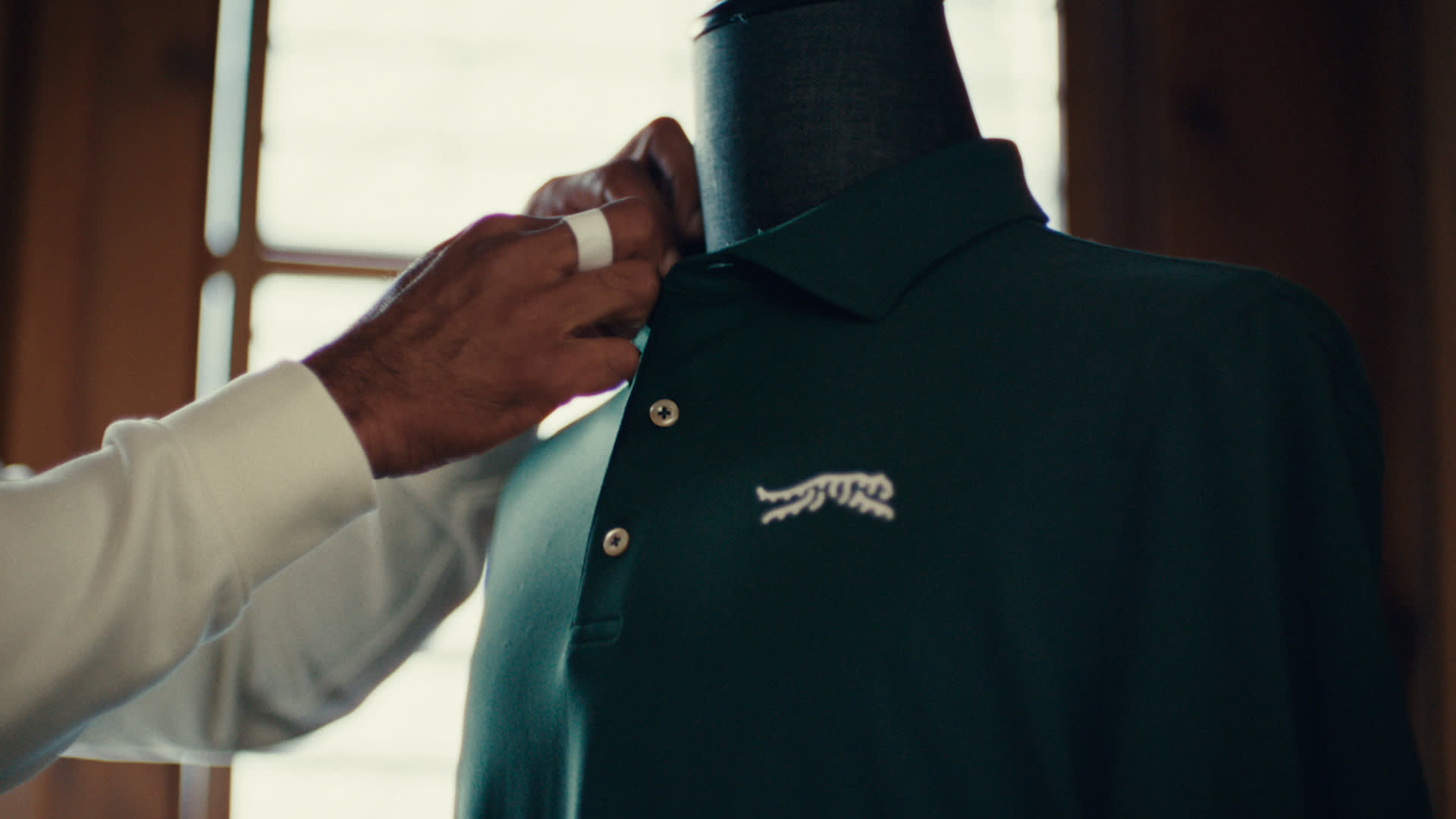 A person adjusts the collar of a dark green polo shirt on a mannequin. The shirt features a white tiger logo on the chest. Light from a window shines in the background.