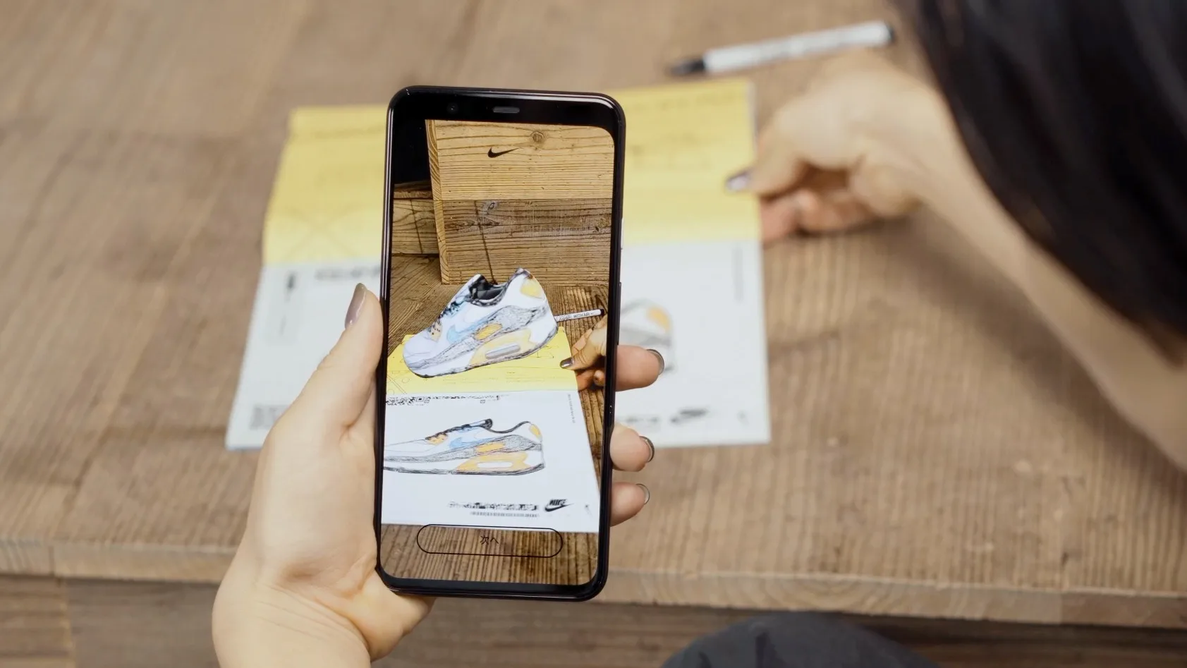 A person uses a smartphone to take a photo of a drawing of a sneaker on a piece of paper, with another hand holding the paper on a wooden surface.