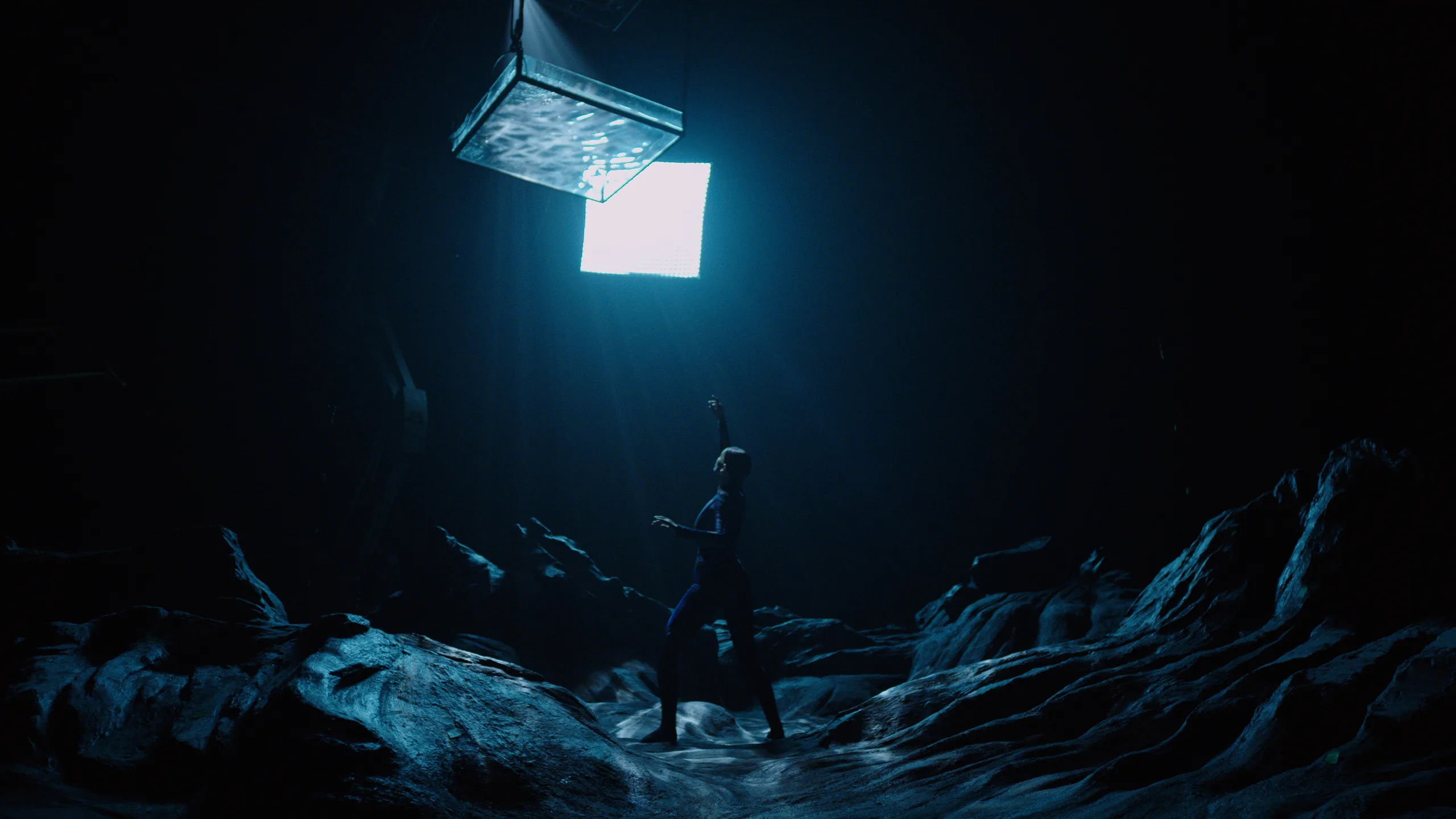 A person stands in a dark, rocky landscape reaching up toward a large, bright, square light source suspended above them, casting dramatic blue light and shadows across the scene.