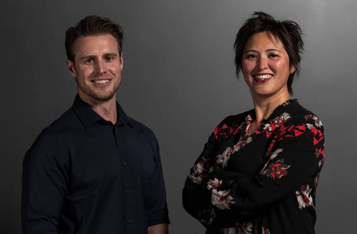 Two people stand side by side against a dark gray background. The man on the left wears a dark shirt and is smiling. The woman on the right is smiling, with arms crossed, wearing a black floral top.