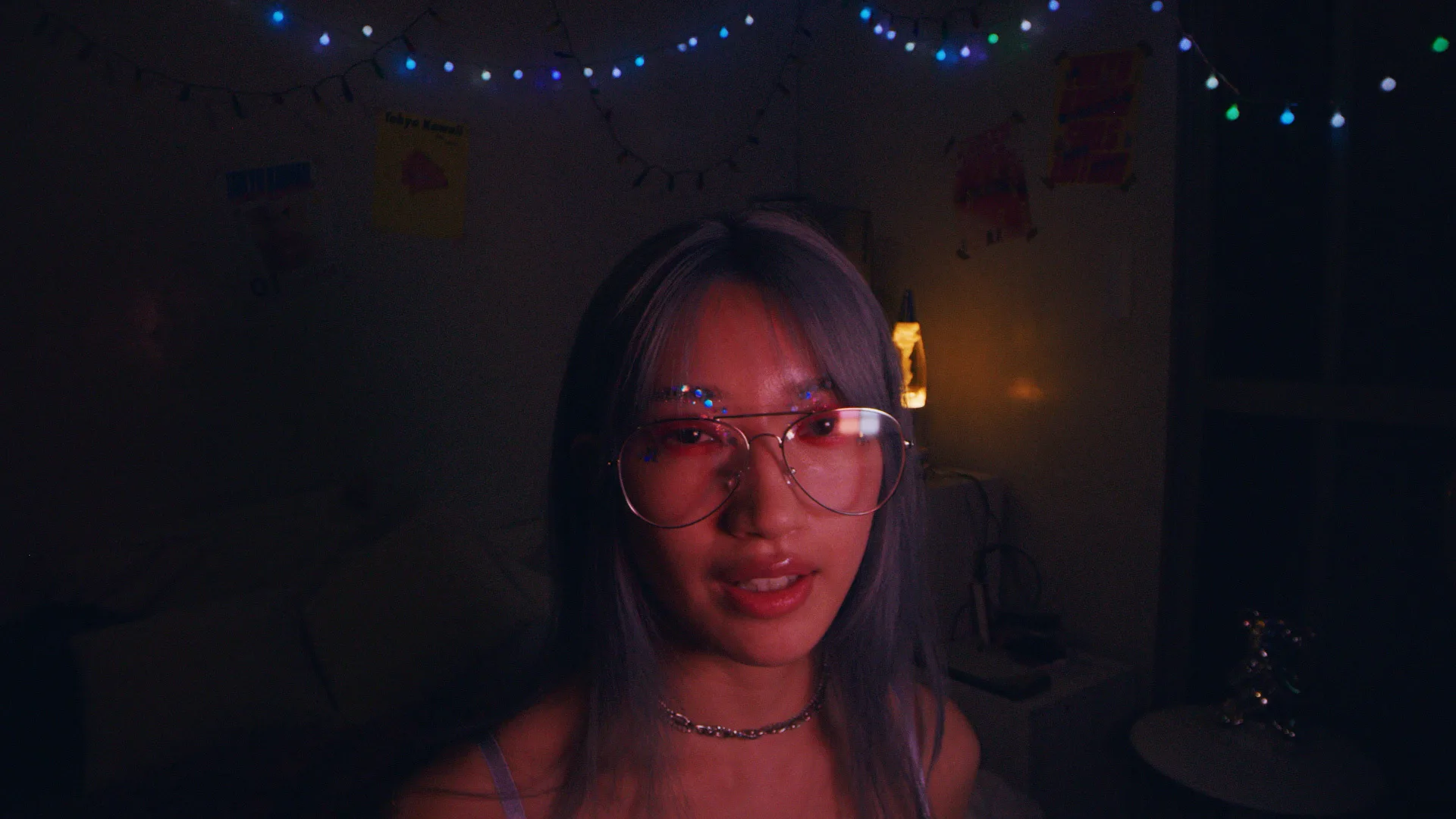 A person with long, blue-tinted hair and large, round glasses sits in a dimly lit room with string lights and posters on the wall. Warm, colorful lighting creates a cozy atmosphere.