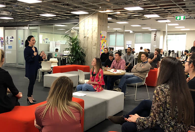 A woman stands and speaks to a group of people seated on couches and chairs in a modern office space with glass walls and open work areas. The audience listens attentively.