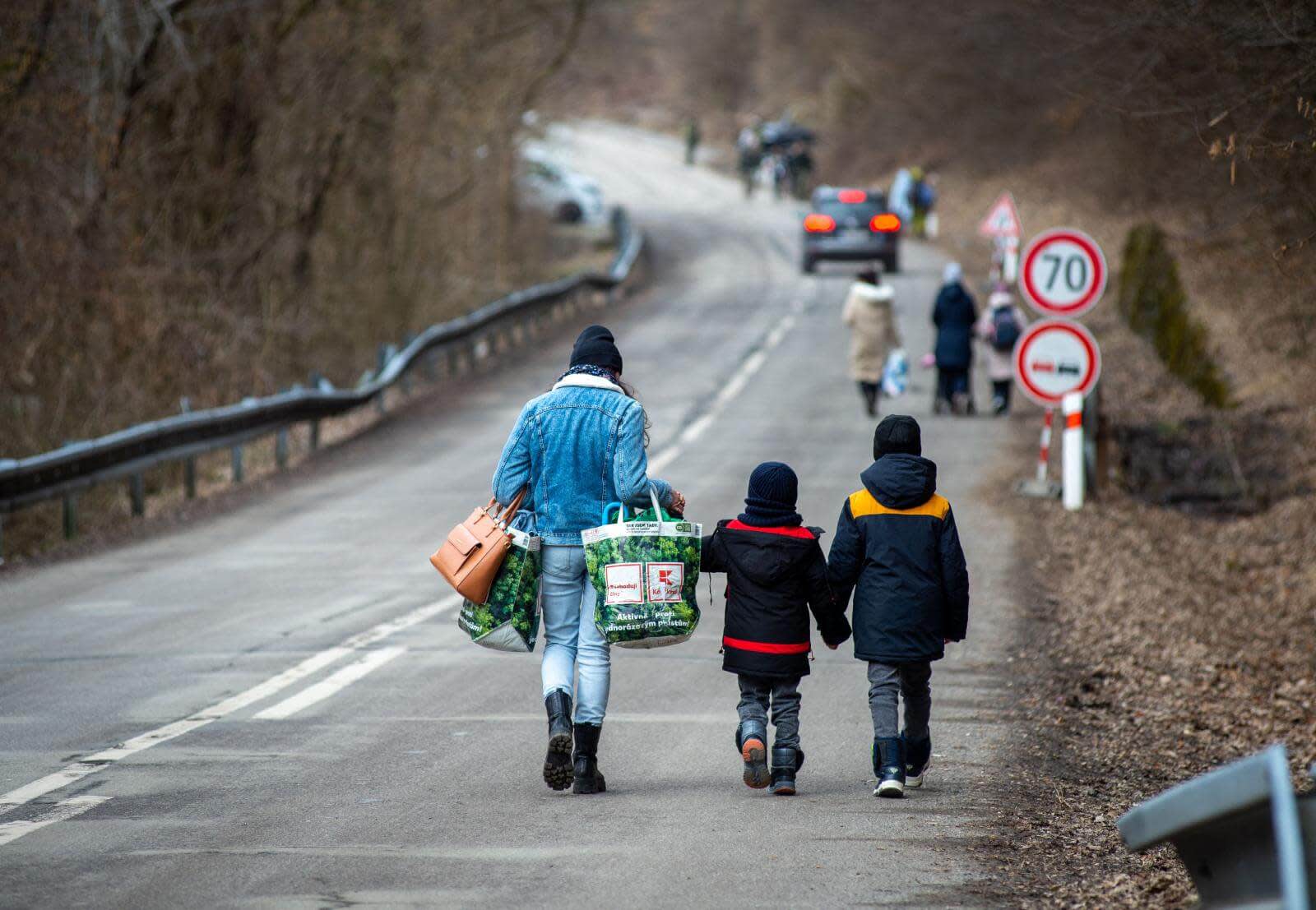 A person carrying bags walks along a rural road with two children, while other people walk ahead. The surroundings are wooded, and speed limit signs are visible on the roadside.