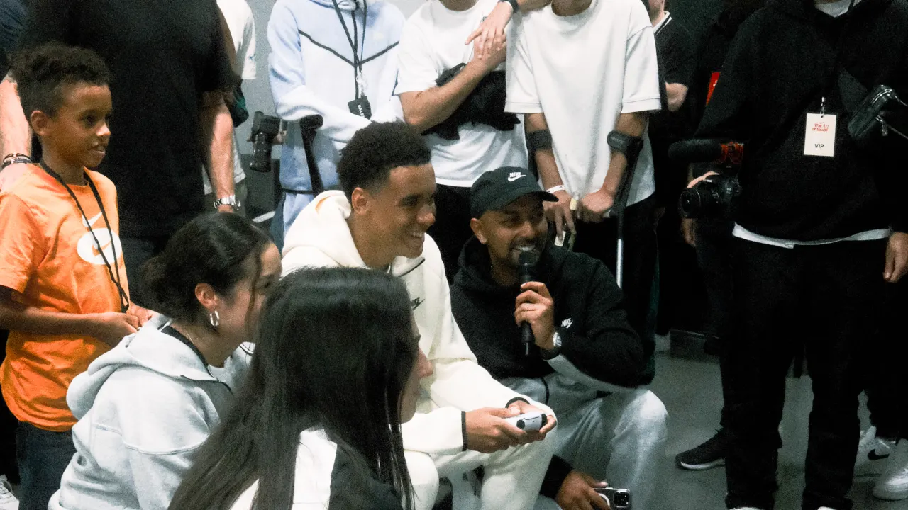 A group of people, including children and adults, gather indoors. Two men, one holding a microphone and the other holding a game controller, are seated at the center, while others stand and watch, smiling and interacting.