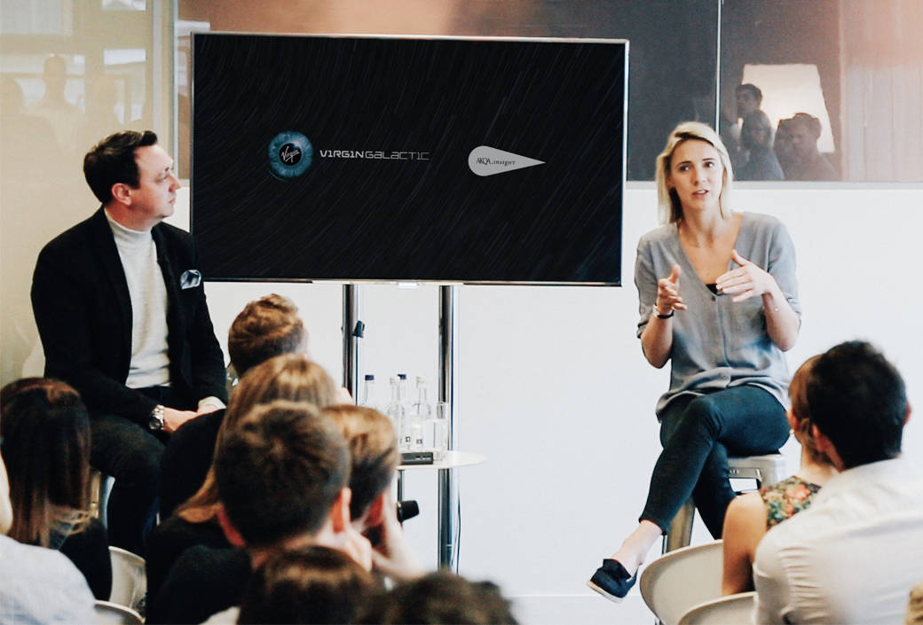 A woman speaks while seated on a stool next to a man during a presentation, with an audience listening and a screen displaying the Virgin Galactic and XL Airways logos behind them.