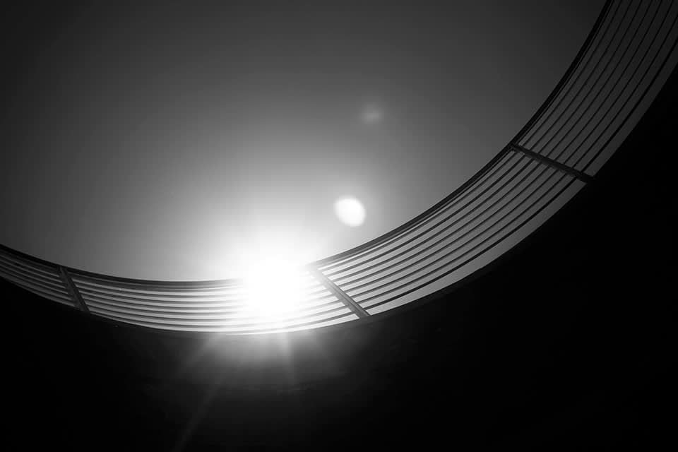 Black and white photo of sunlight shining through a curved metal railing, creating a dramatic silhouette with flares against the sky.