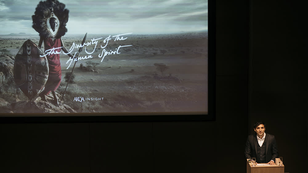 A person in formal attire stands at a podium, presenting in front of a large screen displaying an image of a Maasai warrior and the text The Diversity of the Human Spirit, AKQA Insight.