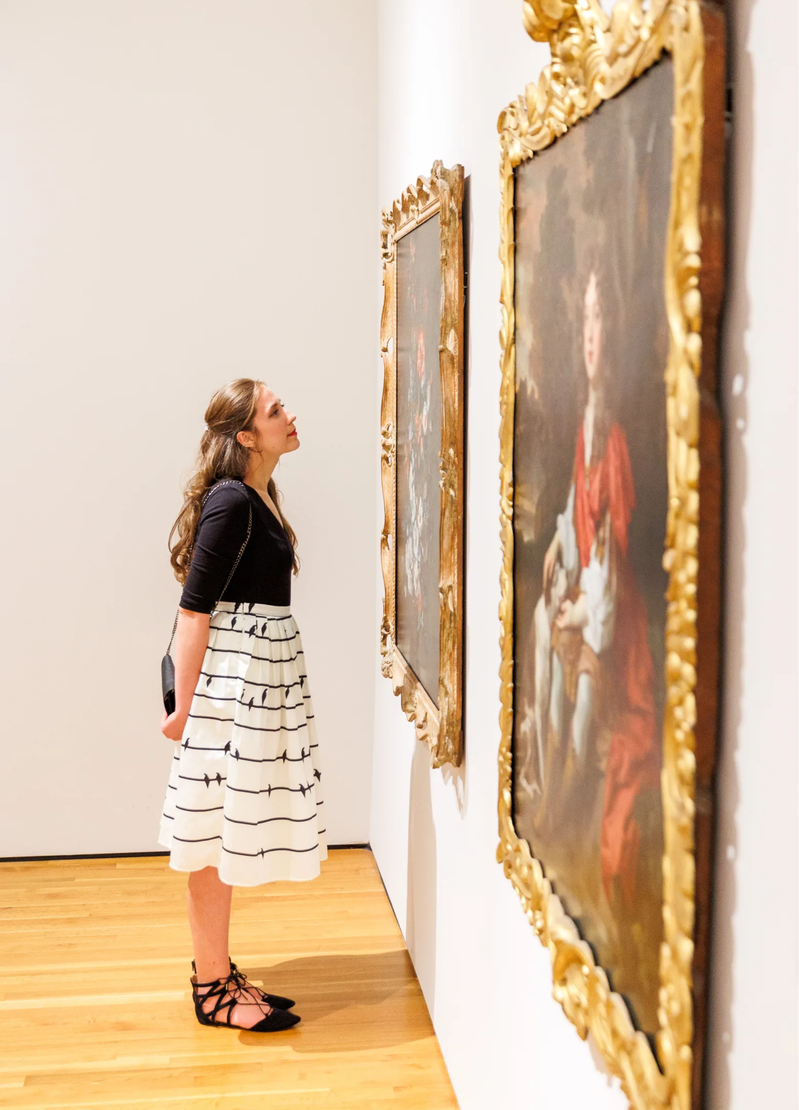A woman in a black top and white skirt stands in an art gallery, closely observing a framed painting on the wall. She holds a clutch and is surrounded by bright, minimalistic surroundings.