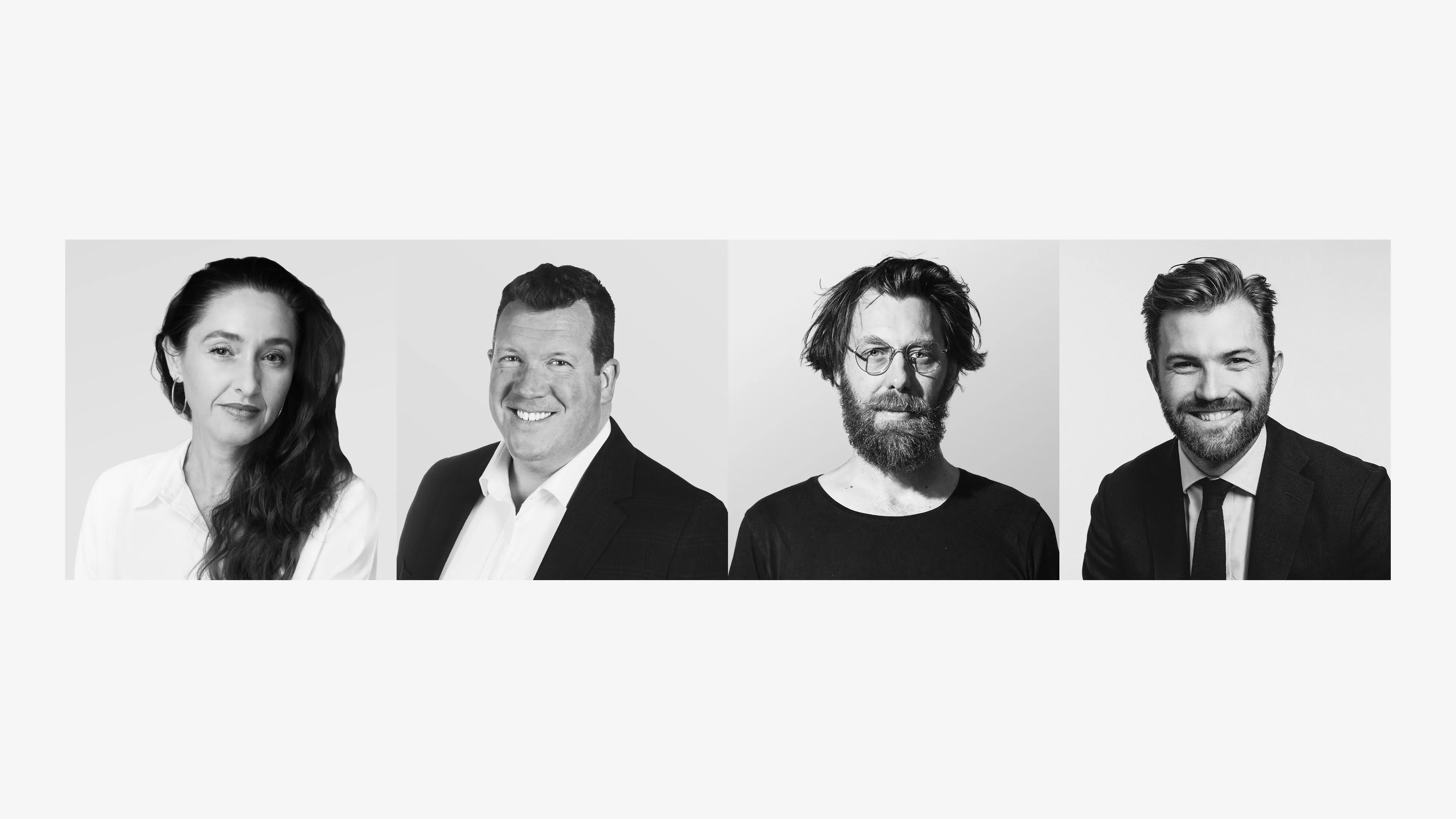 Four people are shown in a row, facing the camera against a plain background. The group includes two men in suits, one man in a casual shirt with glasses and a beard, and one woman with long hair wearing a white blouse.