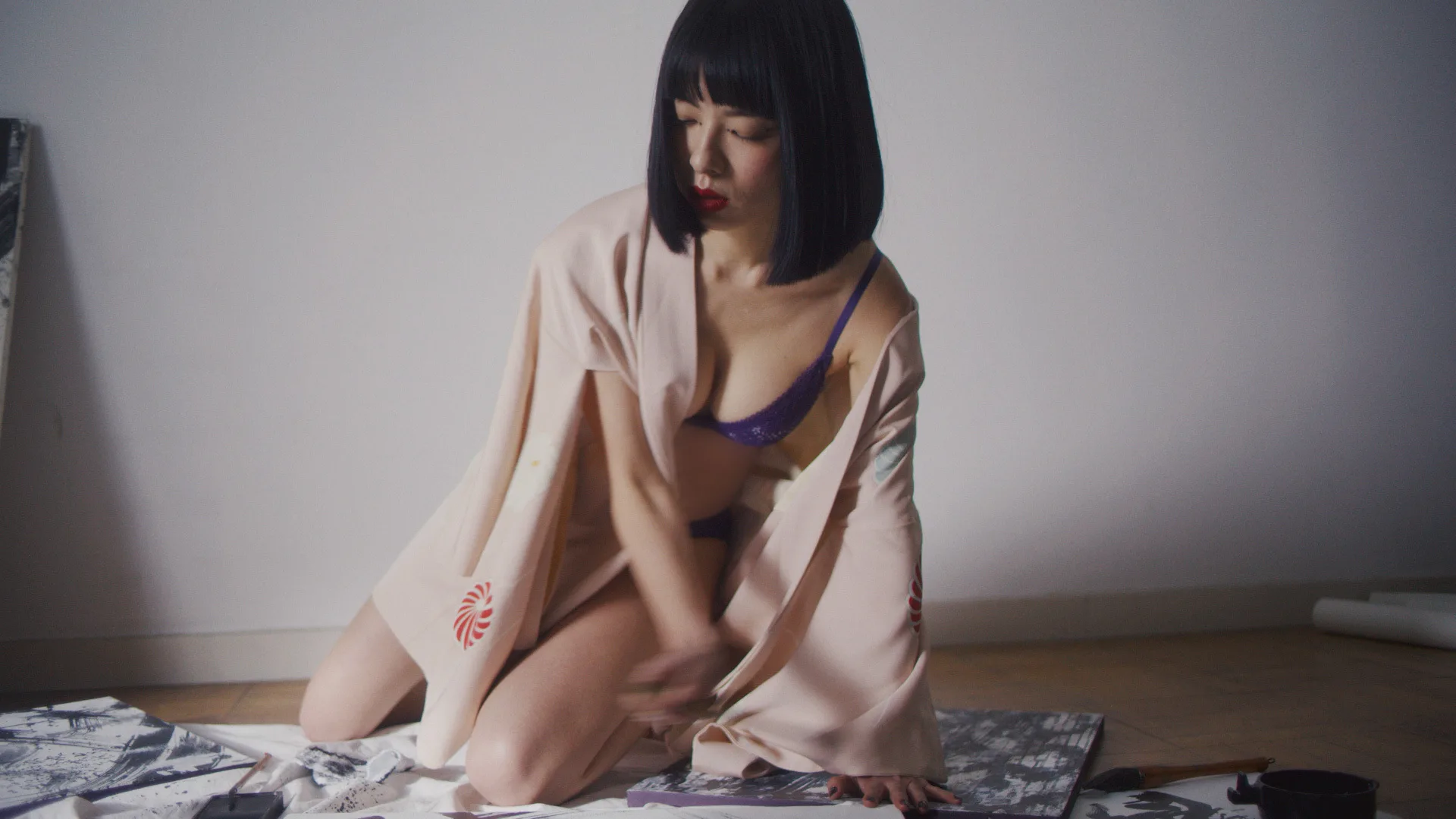 A woman with a bob haircut, wearing a light robe over purple lingerie, kneels on the floor, surrounded by sheets of paper with artwork, painting or drawing with focused expression.