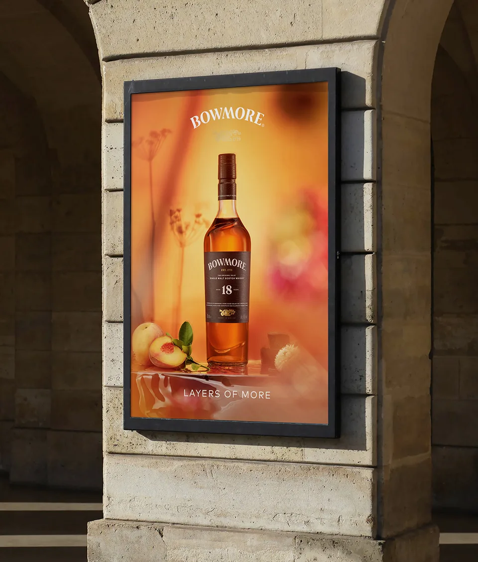 A framed outdoor advertisement displays a bottle of Bowmore 18-year-old whisky alongside sliced peaches, with the tagline LAYERS OF MORE against a warm, orange background.