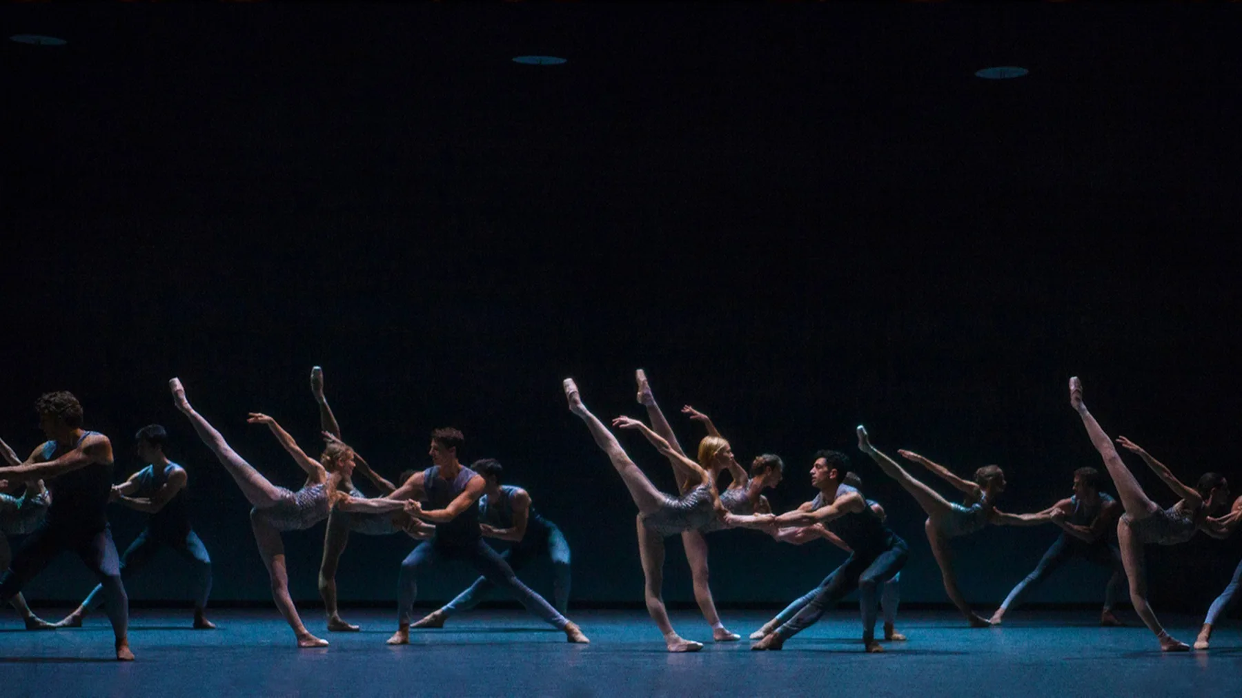 A group of ballet dancers perform on stage in pairs, each female dancer raising one leg high while supported by a male partner. The stage is dimly lit, highlighting their synchronized movements and graceful poses.