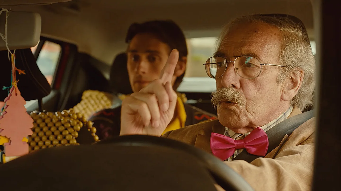 An elderly man with glasses and a pink bow tie gestures while driving a car, with a younger man sitting in the back seat. The car interior is decorated with colorful hanging ornaments.