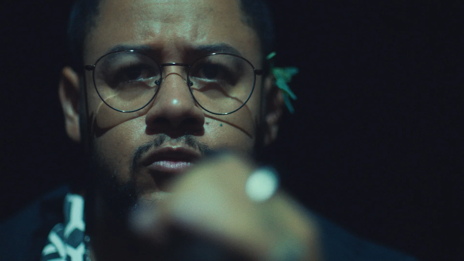 A person with a beard and glasses looks into the camera. They wear a patterned shirt and have a small green plant tucked behind their ear. The image is dimly lit, and their hand is raised in the foreground, slightly out of focus.